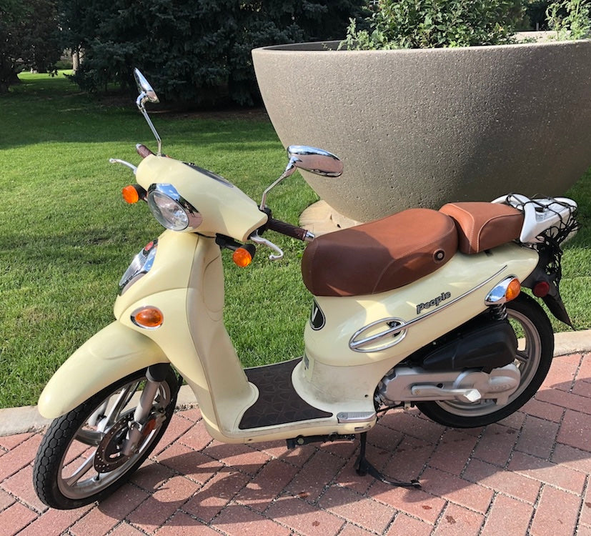 Cream Kymco People 50 scooter outdoors with a cinnamon brown two-piece seat cover set installed, full side view on brick paving with the People branding visible
