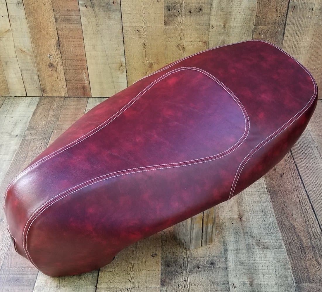 Three-quarter front view of an oxblood Kymco Like seat cover on rustic wood, showing the elongated shape and cream contrast stitching, handmade by Cheeky Seats