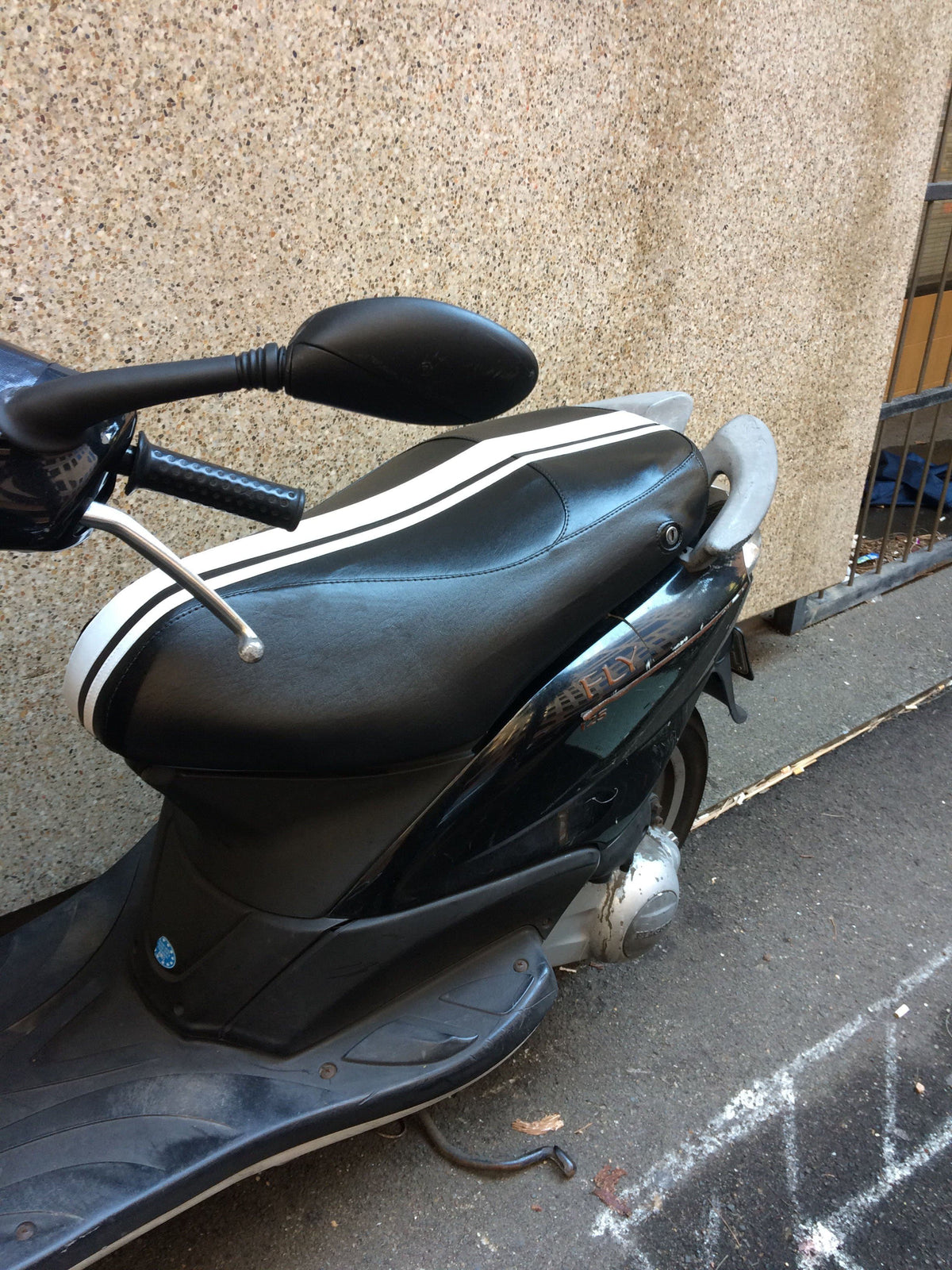 Close installed view of a black Piaggio Fly seat cover with two white racing stripes and visible lock opening, handmade by Cheeky Seats