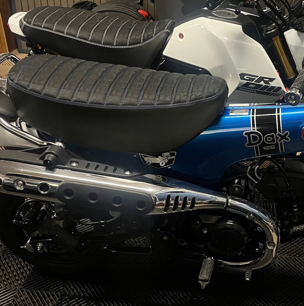 Blue Honda DAX 125 fitted with a black RECARO tuck-and-roll seat cover beside a Honda Grom, with both seats handmade by Cheeky Seats