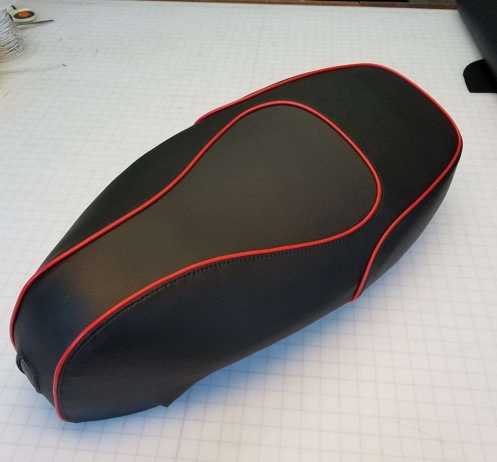 Three-quarter rear view on a cutting mat showing a black Kymco Like seat cover with bold red piping along all seams and the lock opening visible, handmade by Cheeky Seats