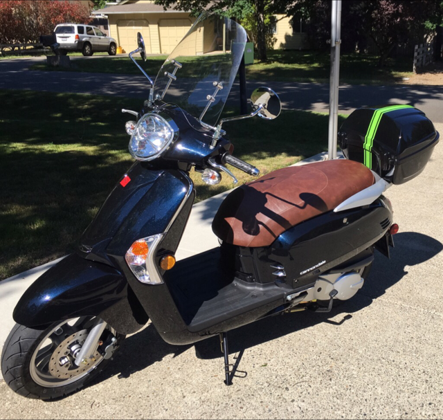 Customer photo of a black Kymco Like scooter outdoors with the whiskey brown seat cover installed, windshield and rear top case visible