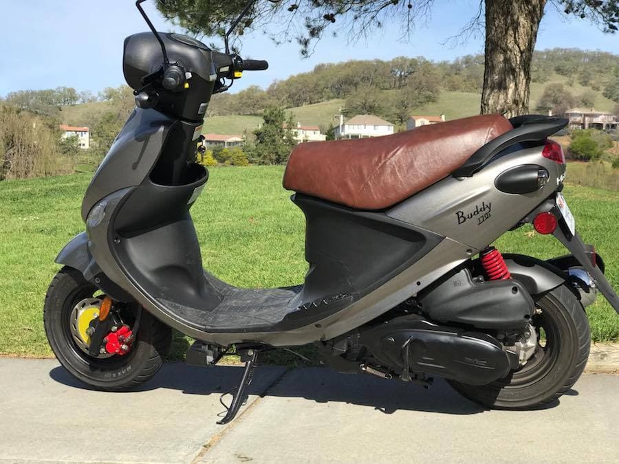 Distressed whiskey-brown Cheeky Seats cover on a titanium Genuine Buddy 125, left-side view